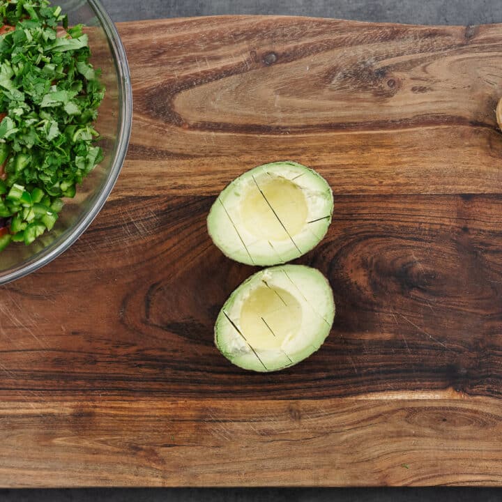 A halved avocado with the creamy flesh sliced into neat lines on a wooden cutting board.