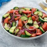 Fresh Mediterranean cucumber and tomato salad served in a white bowl on a white table. A blue towel, a golden fork, and a small cup of black pepper powder are arranged in the upper corner.