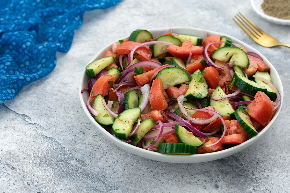 Fresh Mediterranean cucumber tomato salad in a white bowl on a white table with a blue towel, golden fork, and a small cup of black pepper powder nearby.