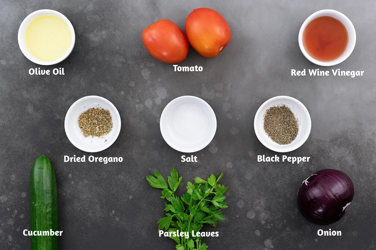 Ingredients for a cucumber tomato salad displayed on a gray table, including olive oil, tomatoes, red wine vinegar, dried oregano, salt, black pepper powder, cucumbers, parsley leaves, and onion.