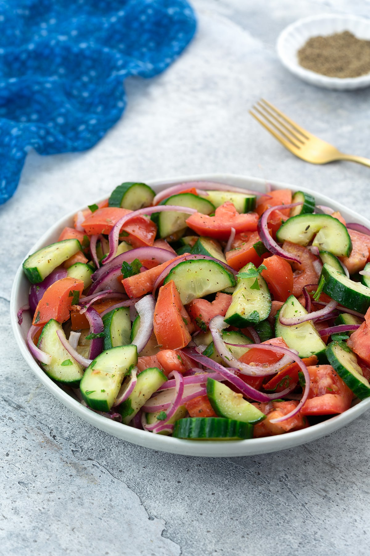 Fresh Mediterranean cucumber tomato salad in a white bowl on a white table with a blue towel, golden fork, and a small cup of black pepper powder nearby.