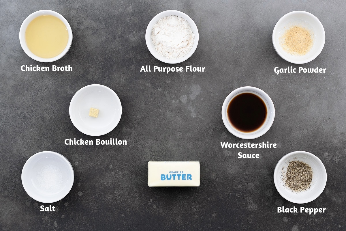 Gravy ingredients arranged on a gray table, including chicken broth, all-purpose flour, garlic powder, chicken bouillon, Worcestershire sauce, salt, butter, and black pepper.