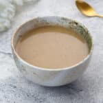 Homemade gravy in a white and brown patterned bowl on a white table, with a towel and a golden spoon placed nearby.