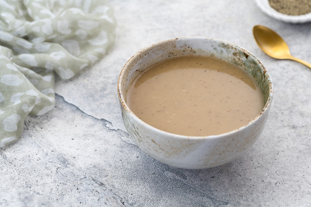 Homemade gravy in a white and brown patterned bowl on a white table. A towel, a small plate of black pepper, and a golden spoon are placed nearby.