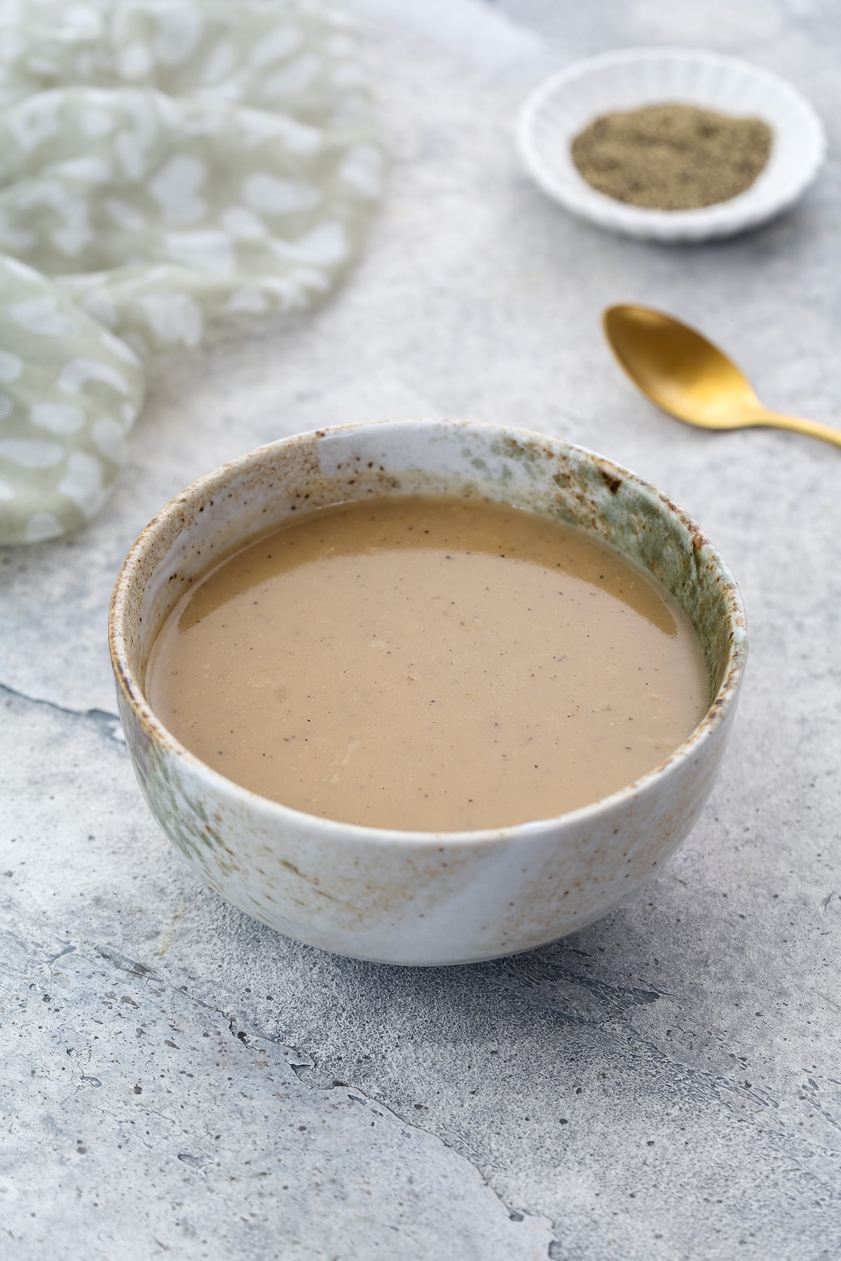 Homemade gravy in a white and brown patterned bowl on a white table. A towel, a small plate of black pepper, and a golden spoon are placed nearby.