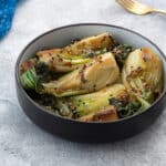 A bowl of cooked bok choy in a brown bowl on a gray table, surrounded by a blue towel, and a golden fork.