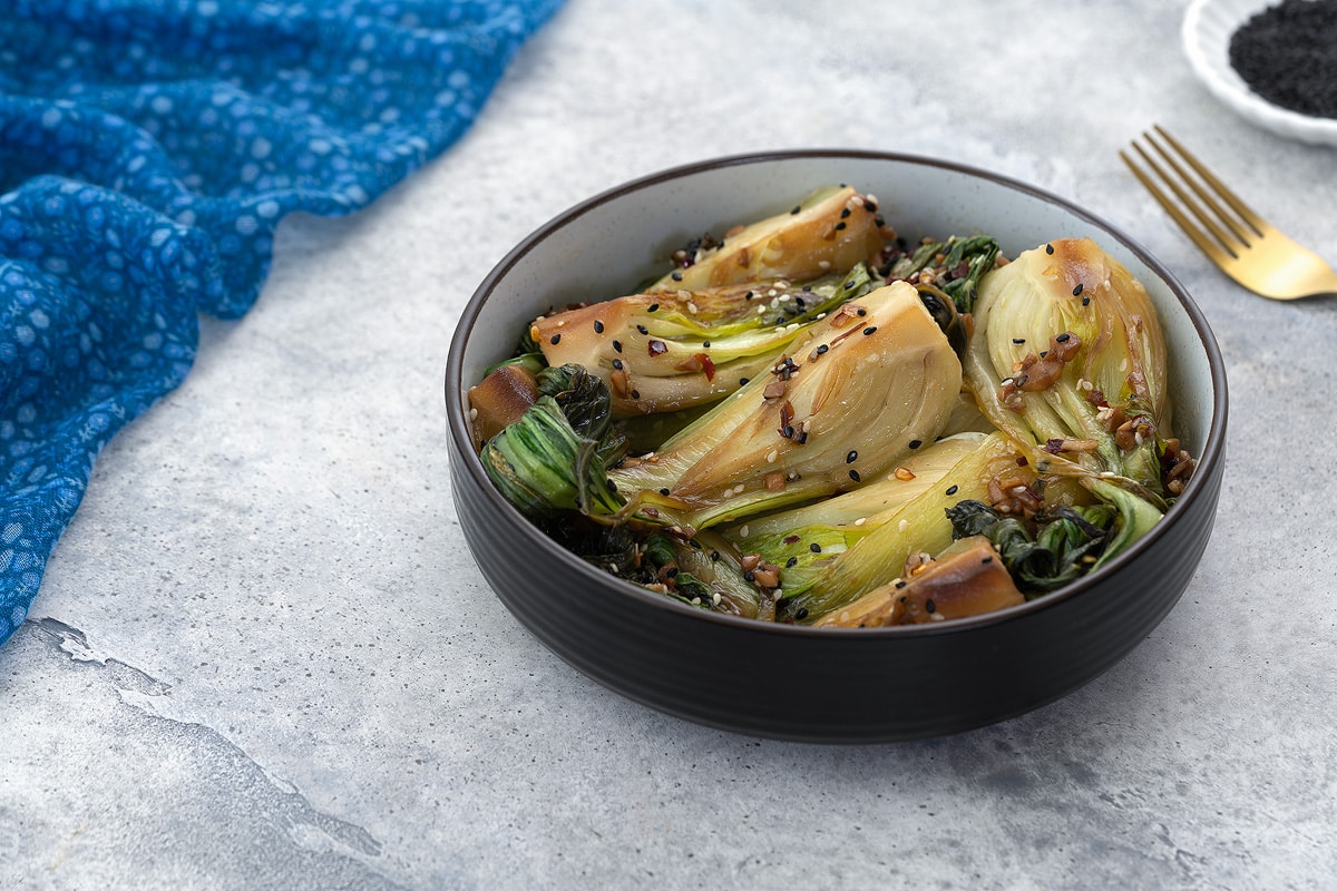 A bowl of cooked bok choy in a brown bowl on a gray table, surrounded by a blue towel, a small plate of black sesame seeds, and a golden fork.