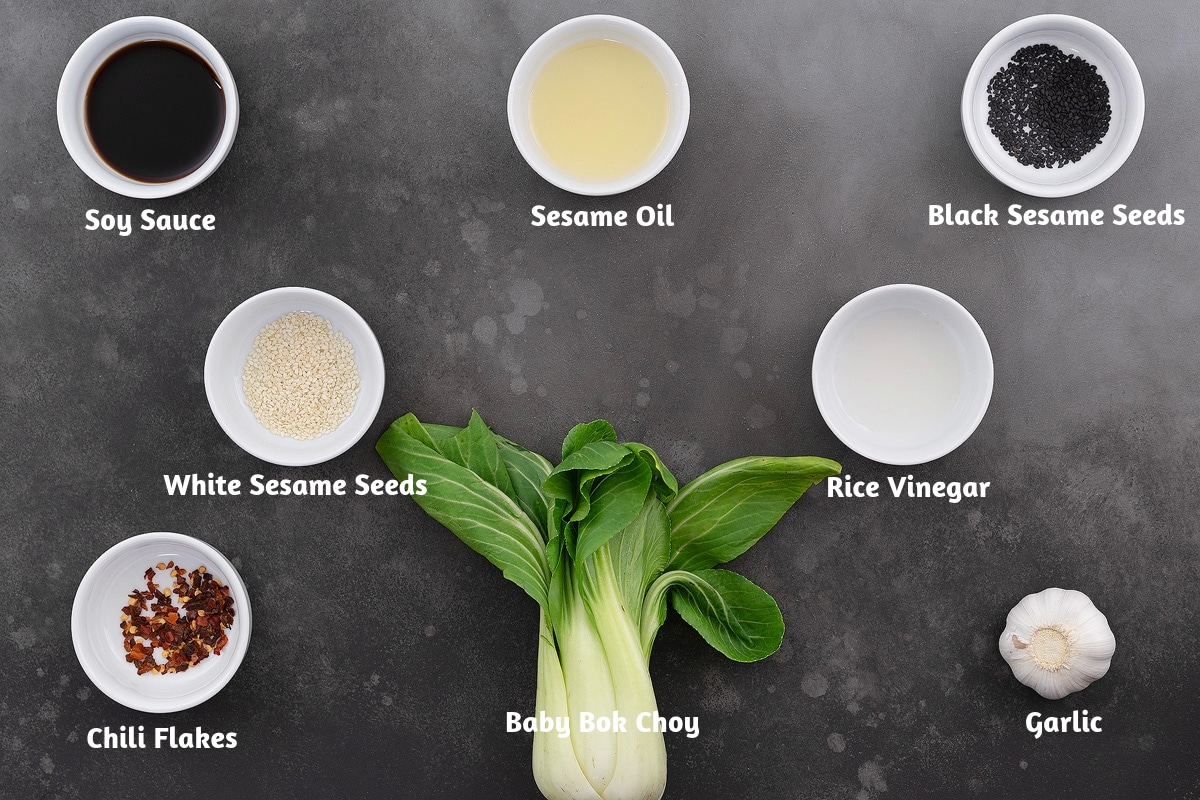 Ingredients for sautéed bok choy on a gray table: soy sauce, sesame oil, black sesame seeds, white sesame seeds, rice vinegar, chili flakes, baby bok choy, and garlic.