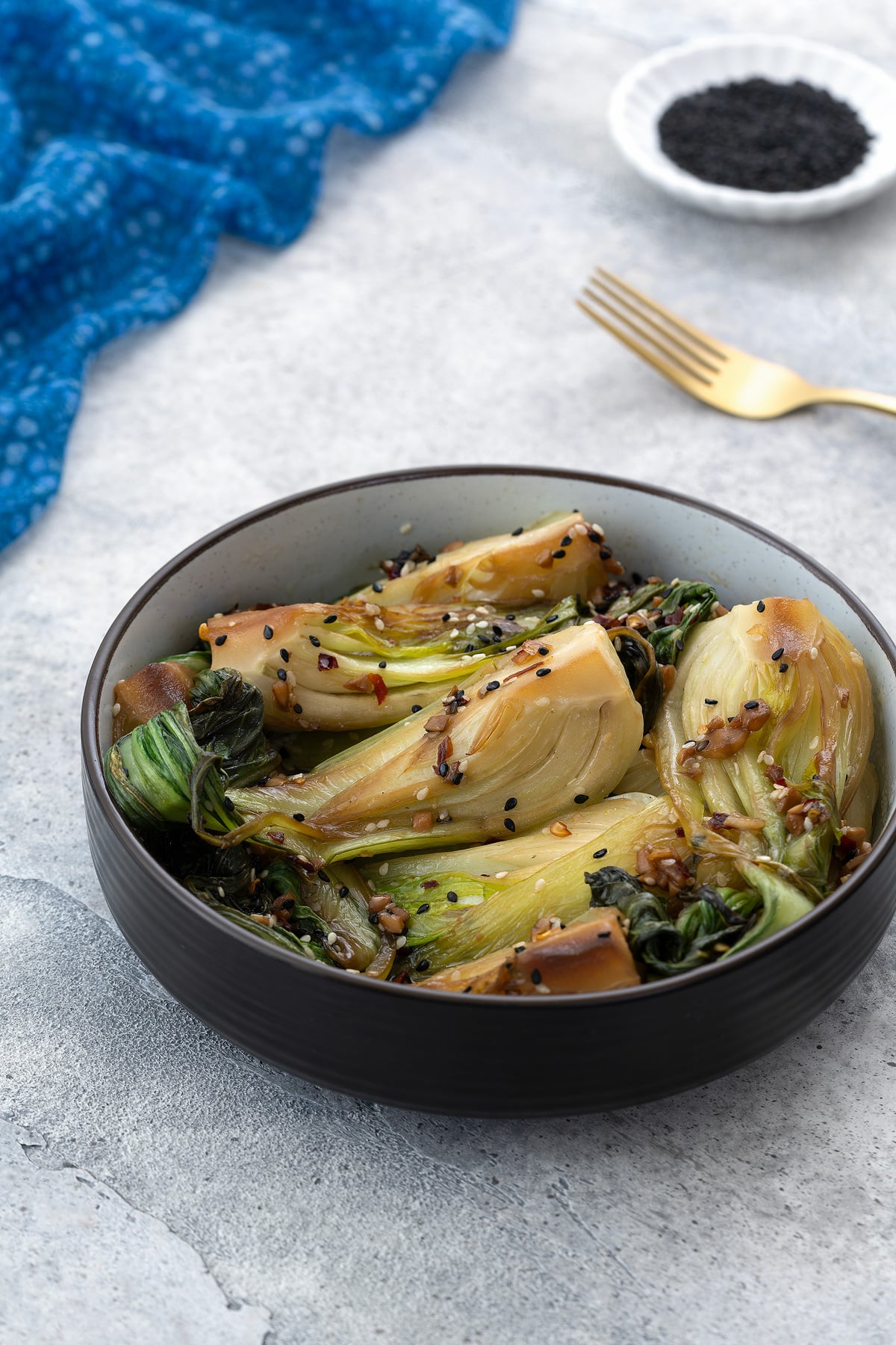 A bowl of cooked bok choy in a brown bowl on a gray table, surrounded by a blue towel, a small plate of black sesame seeds, and a golden fork.