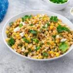 Homemade Mexican corn salad in a white bowl on a white table. A blue towel and a cup of sliced scallions are arranged in the top corner.