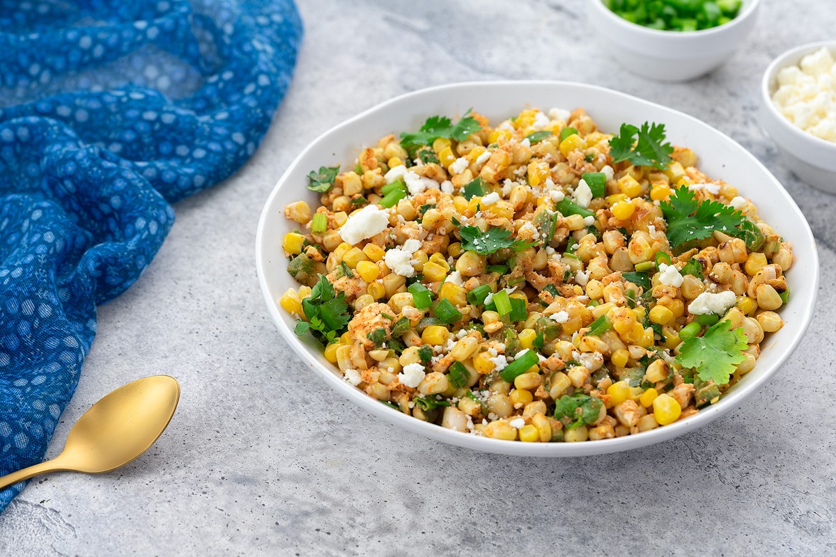 Homemade Mexican corn salad in a white bowl on a white table. A blue towel, a cup of sliced scallions, and feta cheese are arranged nearby.