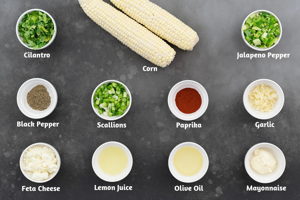 Mexican corn salad ingredients on a gray table, including cilantro, corn, jalapeno pepper, black pepper, scallions, paprika, garlic, feta cheese, lemon juice, olive oil, and mayonnaise.