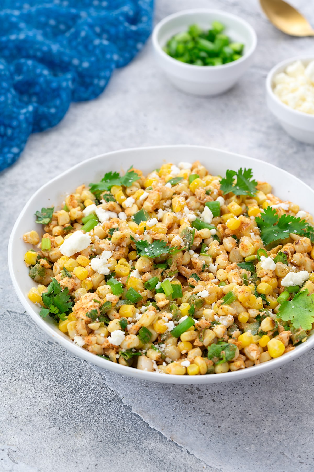 Homemade Mexican corn salad in a white bowl on a white table. A blue towel, a cup of sliced scallions, and feta cheese are arranged nearby.