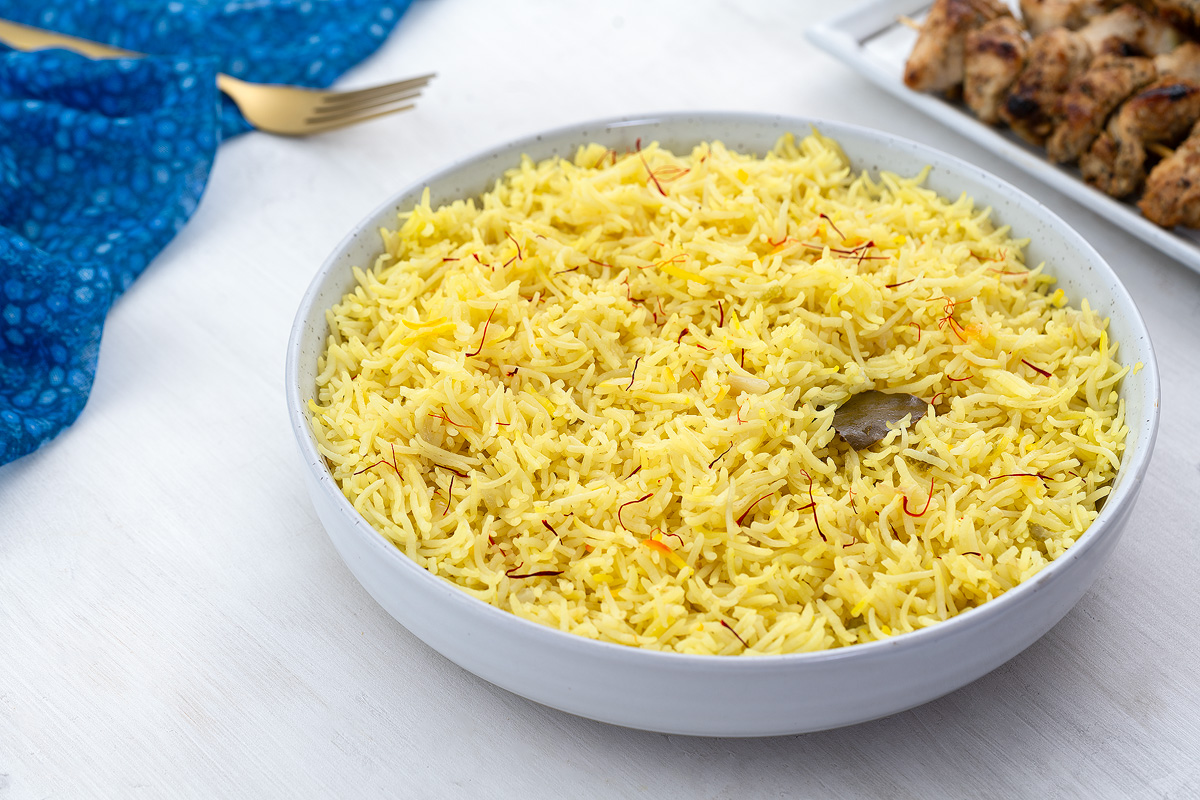 Homemade saffron rice in a white bowl on a white table, surrounded by a blue towel, golden fork, and a plate of chicken kabobs