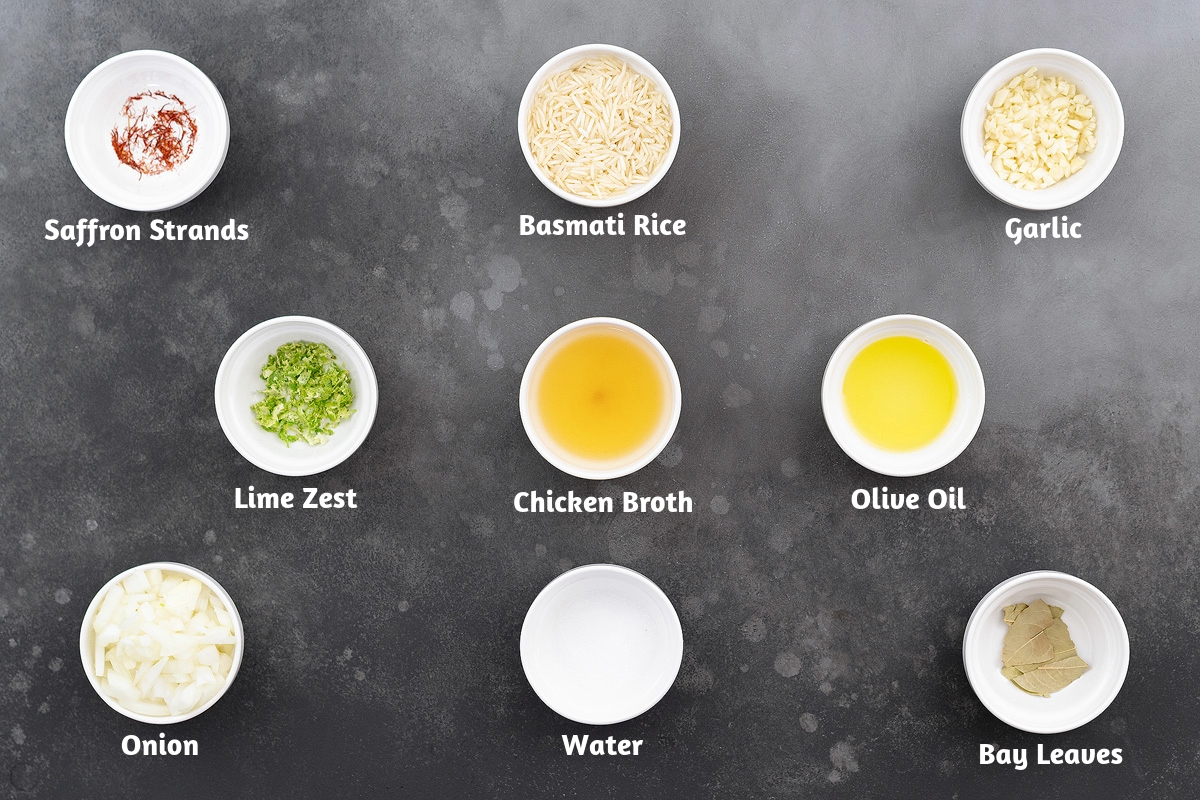 Ingredients for saffron rice arranged on a gray table: saffron strands, basmati rice, garlic, lime zest, chicken broth, olive oil, onion, water, and bay leaves.