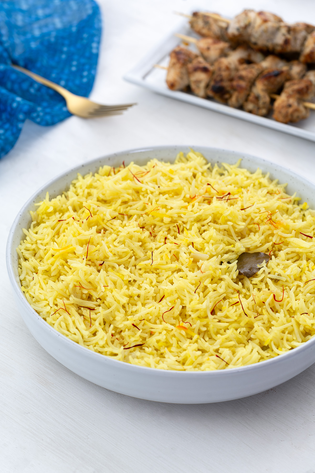 Homemade saffron rice in a white bowl on a white table, surrounded by a blue towel, golden fork, and a plate of chicken kabobs