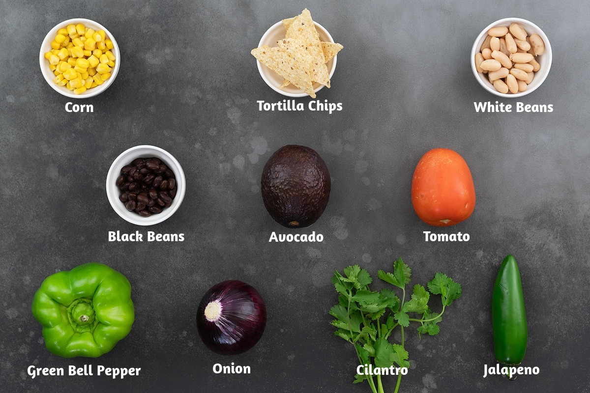 Ingredients for Texas Cowboy Caviar recipe displayed on a gray table, including corn, tortilla chips, white beans, black beans, avocado, tomato, green bell pepper, onion, cilantro, and jalapeno.