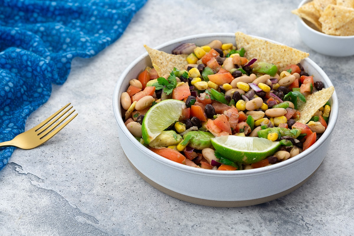 Homemade Texas Cowboy Caviar served in a white bowl on a white table, accompanied by a blue towel, a golden fork, and a cup of tortilla chips nearby.