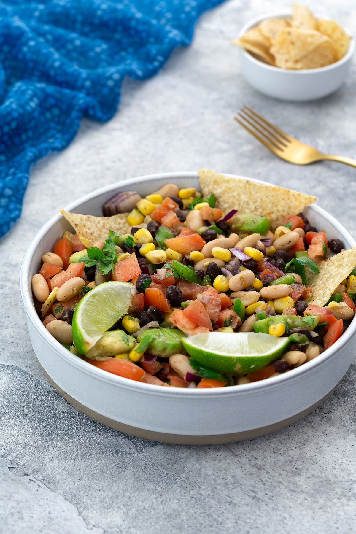 Homemade Texas Cowboy Caviar served in a white bowl on a white table, accompanied by a blue towel, a golden fork, and a cup of tortilla chips nearby.