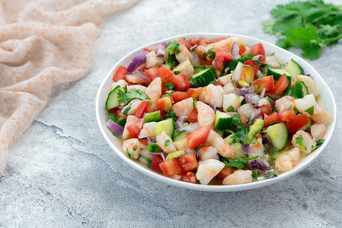 Fresh homemade shrimp ceviche in a white bowl on a white table, with a towel, golden fork, and cilantro nearby.