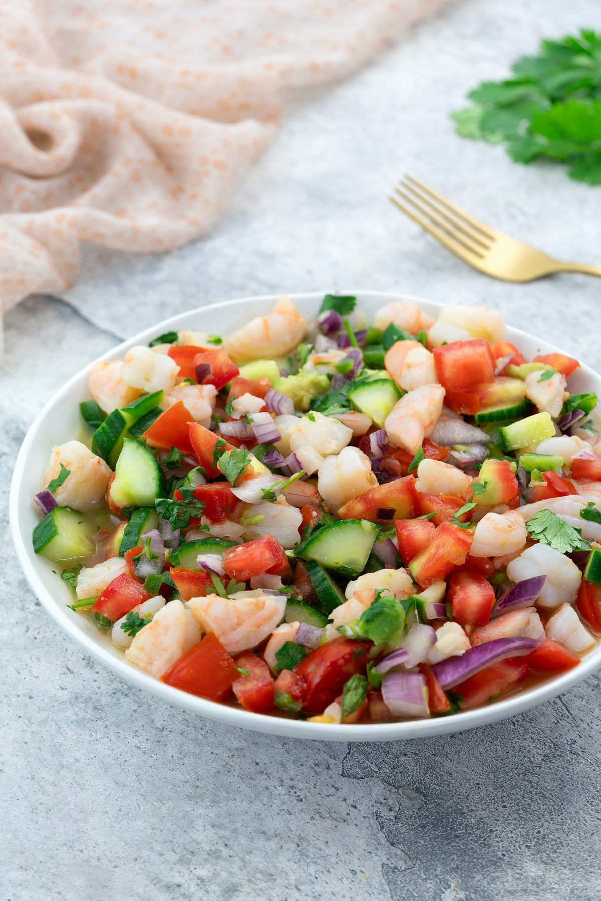 Fresh homemade shrimp ceviche in a white bowl on a white table, with a towel, golden fork, and cilantro nearby.