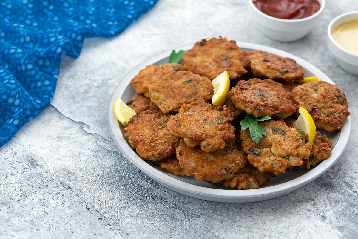 Homemade chicken patties on a white plate, accompanied by lemon wedges, on a white table. Next to them, there are cups of ketchup and honey mustard, with a blue towel arranged nearby.