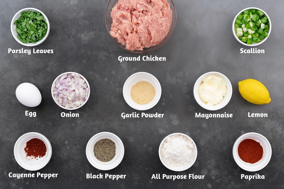 Ingredients for chicken patties arranged on a gray table. The setup includes parsley leaves, ground chicken, scallions, eggs, chopped onion, garlic powder, mayonnaise, lemon, cayenne pepper, black pepper, all-purpose flour, and paprika.