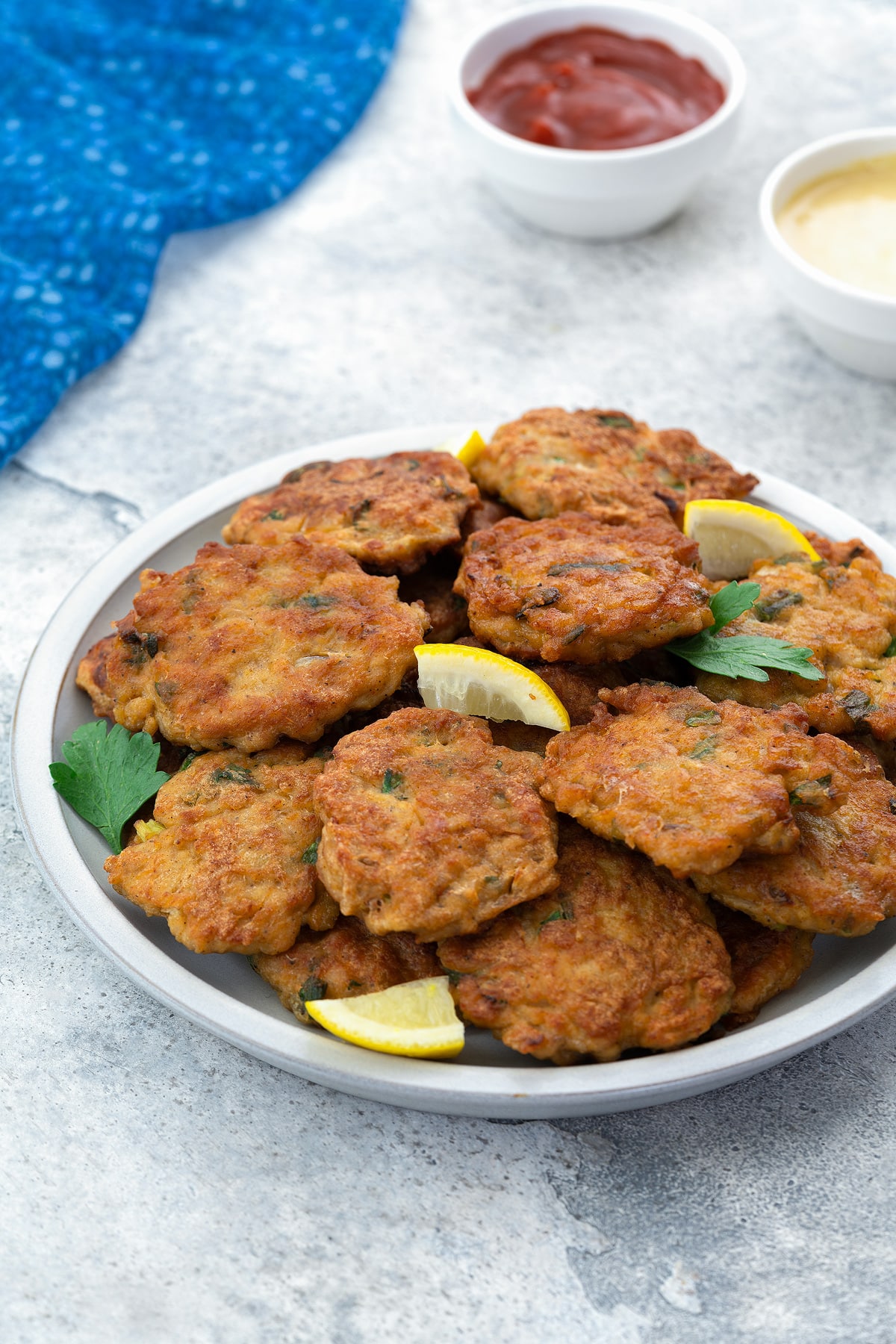 Homemade chicken patties on a white plate, accompanied by lemon wedges, on a white table. Next to them, there are cups of ketchup and honey mustard, with a blue towel arranged nearby.