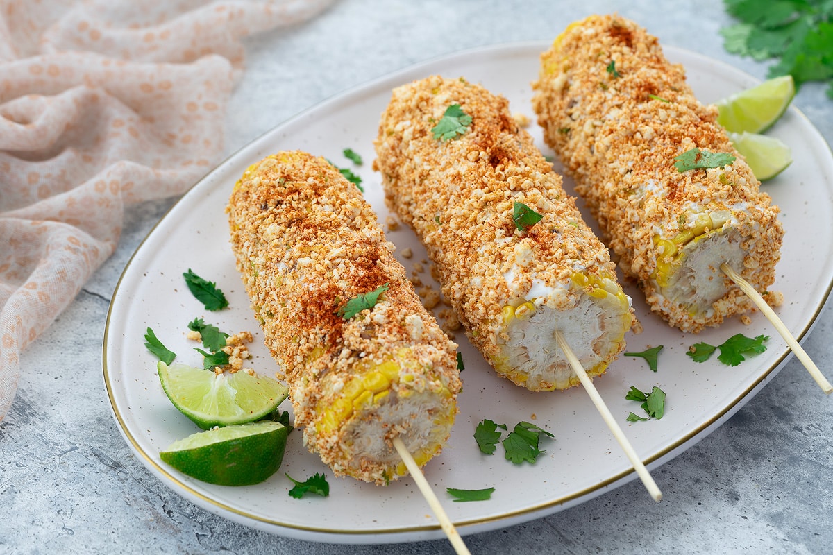 Elote, Mexican street corn, served with lime on an oval plate on a white table. A towel and cilantro placed nearby.