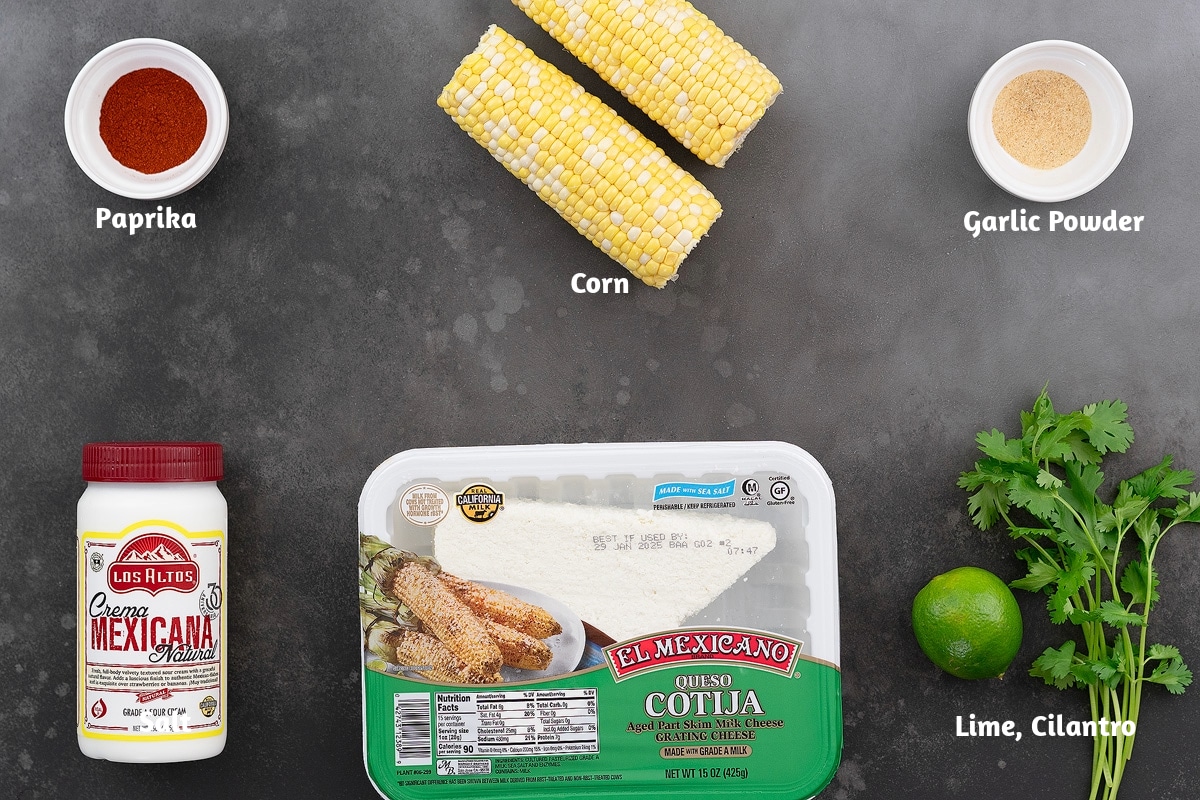 Ingredients for Elote, Mexican street corn, arranged on a gray table, including paprika, corn, garlic powder, Mexican crema, Cotija cheese, lime, and cilantro.