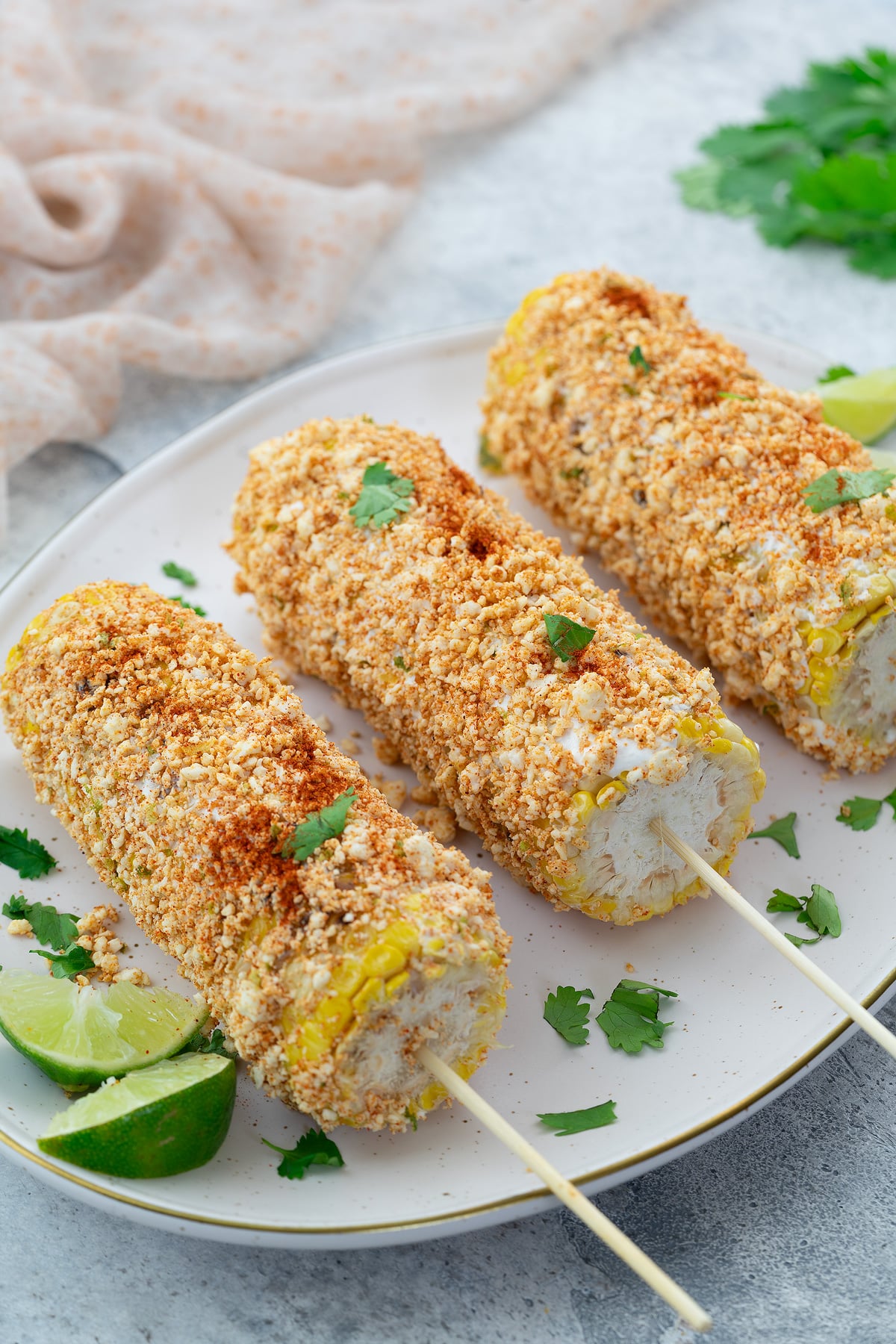 Elote, Mexican street corn, served with lime on an oval plate on a white table. A towel and cilantro placed nearby.