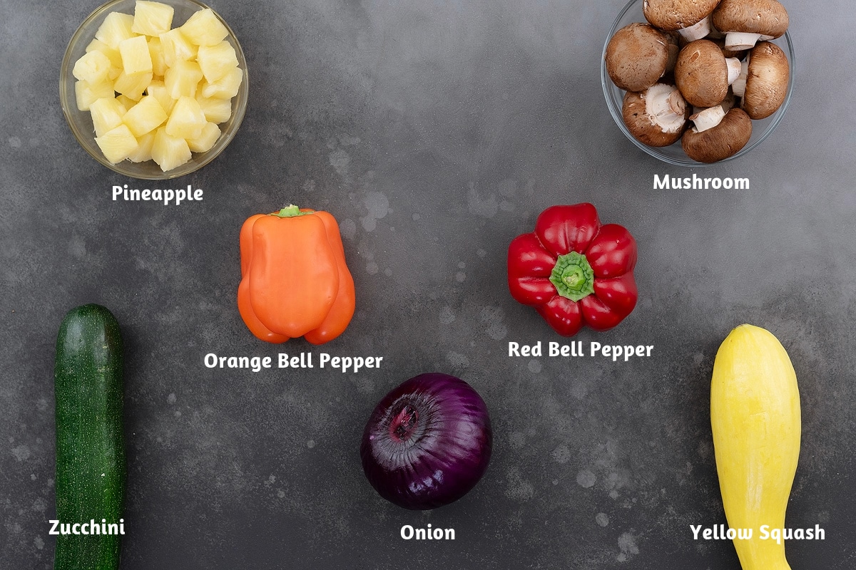 Vegetable skewer ingredients on a gray table, featuring pineapple, mushrooms, orange bell pepper, red bell pepper, zucchini, onion, and yellow squash.