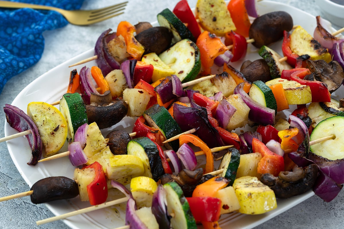 Grilled vegetable skewers on a white plate, surrounded by a blue towel, and a golden fork.