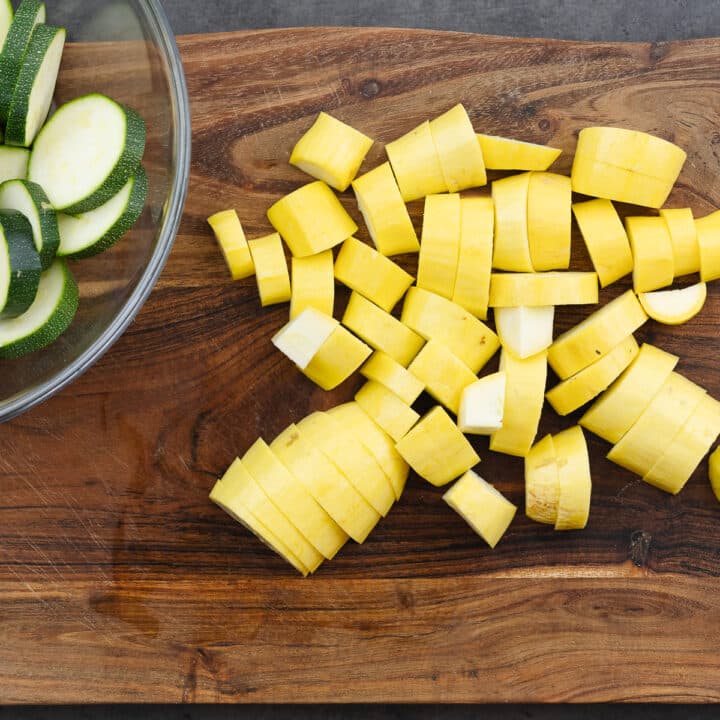 A wooden cutting board with chopped yellow squash, accompanied by a bowl of chopped zucchini on the side.