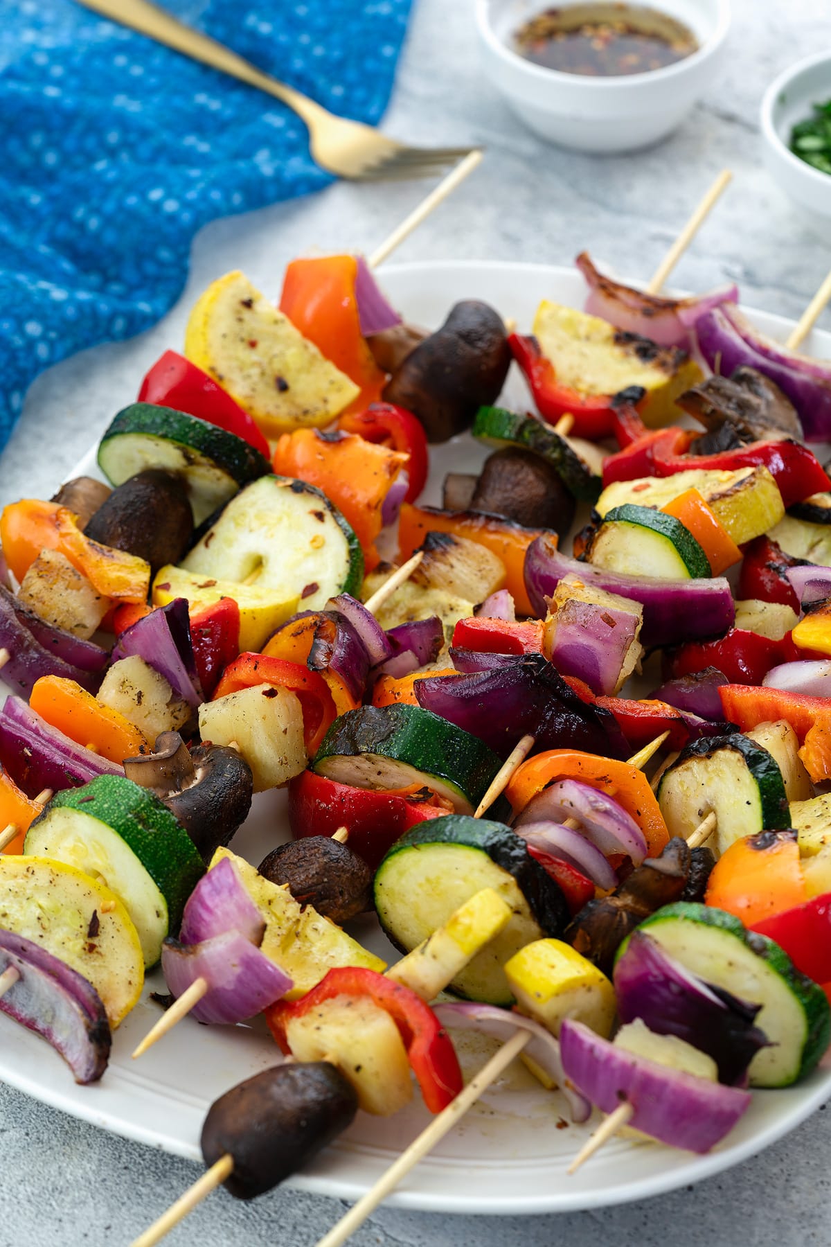 Grilled vegetable skewers on a white plate, surrounded by a blue towel, marinade, golden fork, and chopped cilantro in a small cup, all set on a white table.