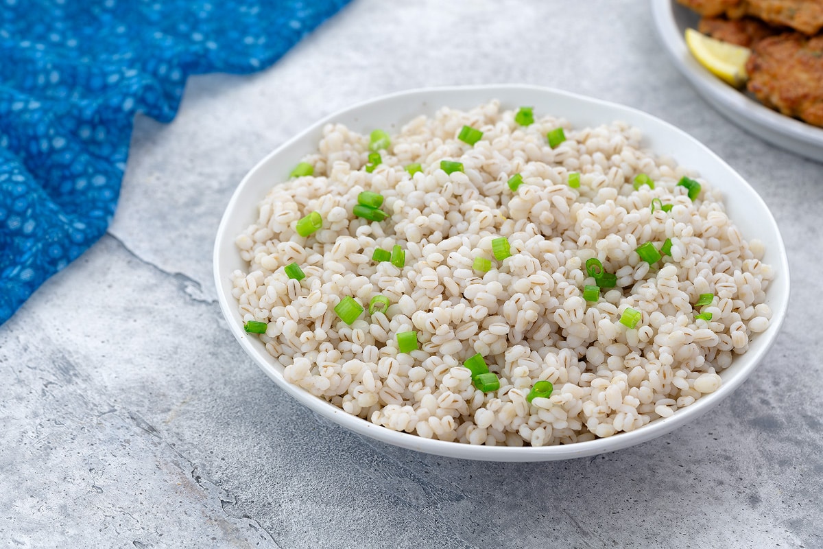 Home-cooked barley in a white bowl on a white table, accompanied by a plate of chicken patties and a blue towel arranged nearby.