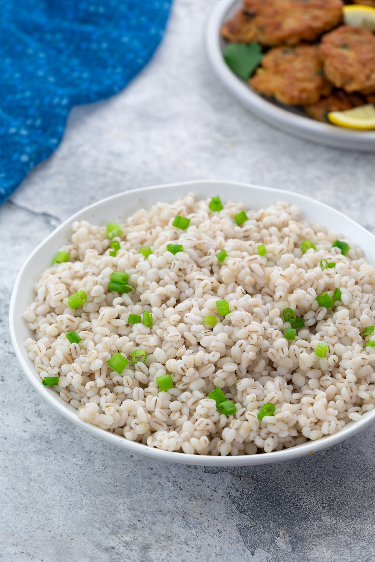 Home-cooked barley in a white bowl on a white table, accompanied by a plate of chicken patties and a blue towel arranged nearby.