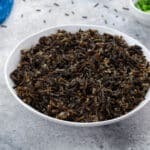 Cooked wild rice in a white bowl on a white table with a blue towel and a small cup of scallions in the top corner.