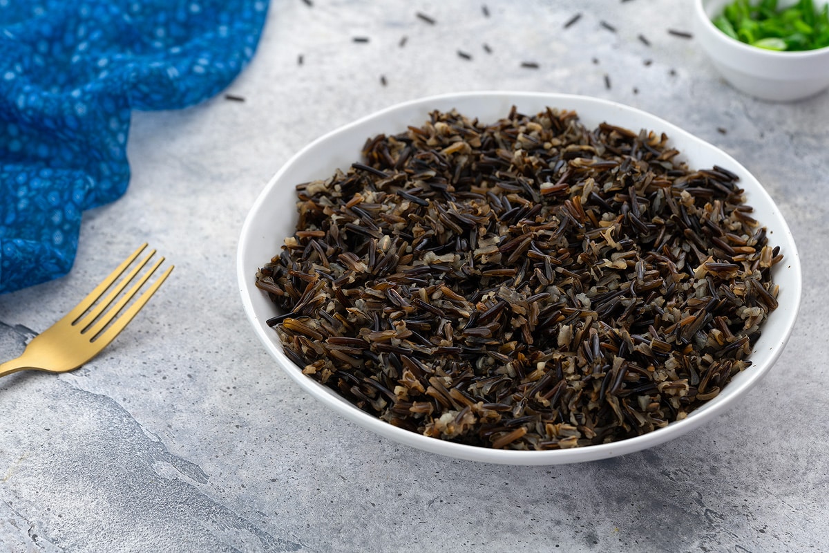 Cooked wild rice in a white bowl on a white table with a blue towel, golden fork, and a small cup of scallions.