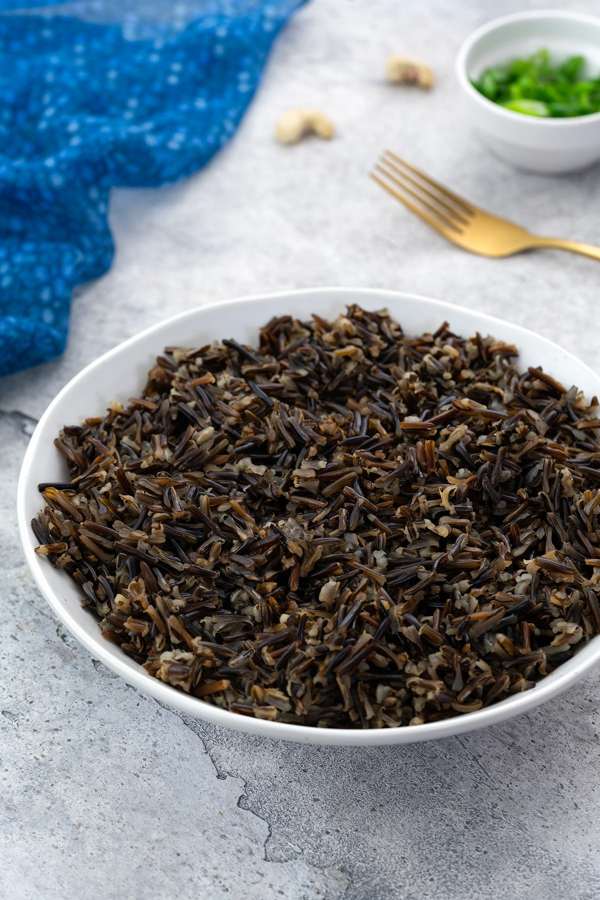 Cooked wild rice in a white bowl on a white table with a blue towel, golden fork, and a small cup of scallions.