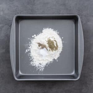 A shallow tray filled with flour, salt, pepper, and Italian seasoning, ready to coat the chicken.