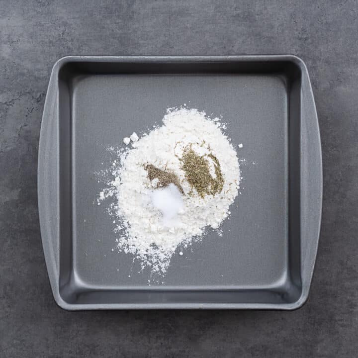 A shallow tray filled with flour, salt, pepper, and Italian seasoning, ready to coat the chicken.