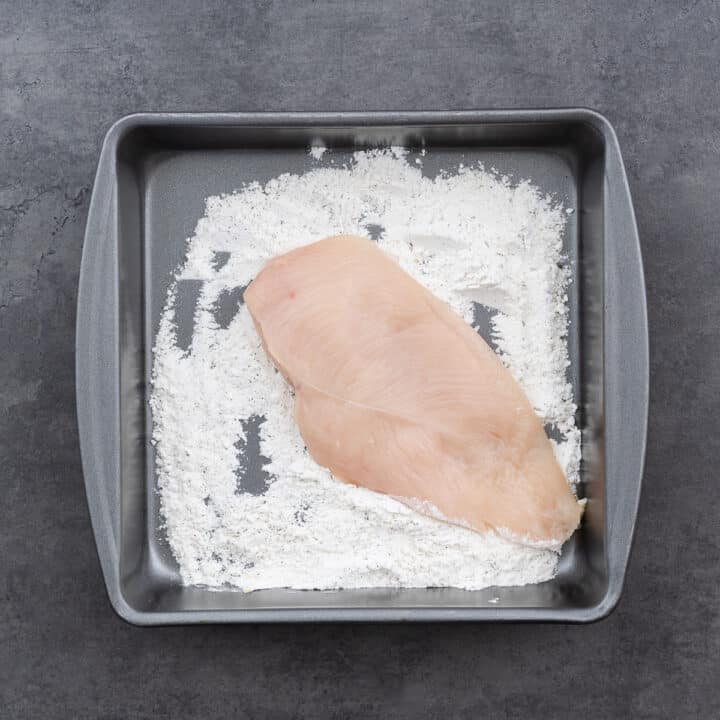 A shallow tray with flour mixture alongside boneless chicken breasts, prepped for coating.