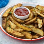 A plate of homemade potato wedges with a cup of ketchup on a white table. A blue towel is in the top left corner.