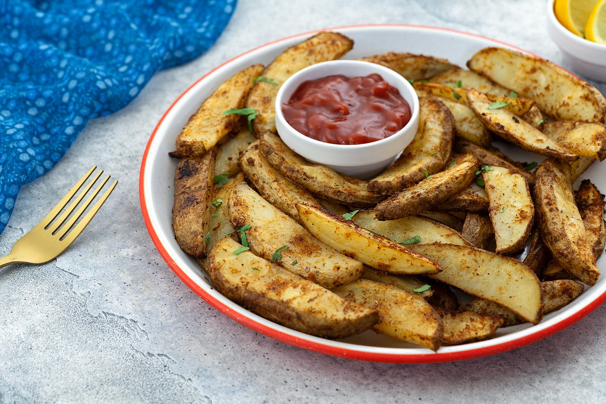 A plate of homemade potato wedges with a cup of ketchup on a white table. A blue towel, golden fork, and a small cup of lemon wedges are placed nearby.