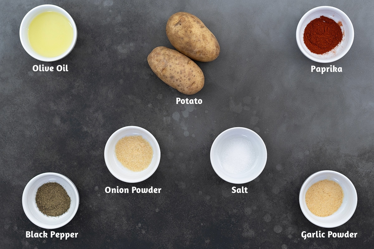 Baked Potato Wedges ingredients placed on a gray table, including olive oil, potato, paprika, onion powder, salt, black pepper, and garlic powder.