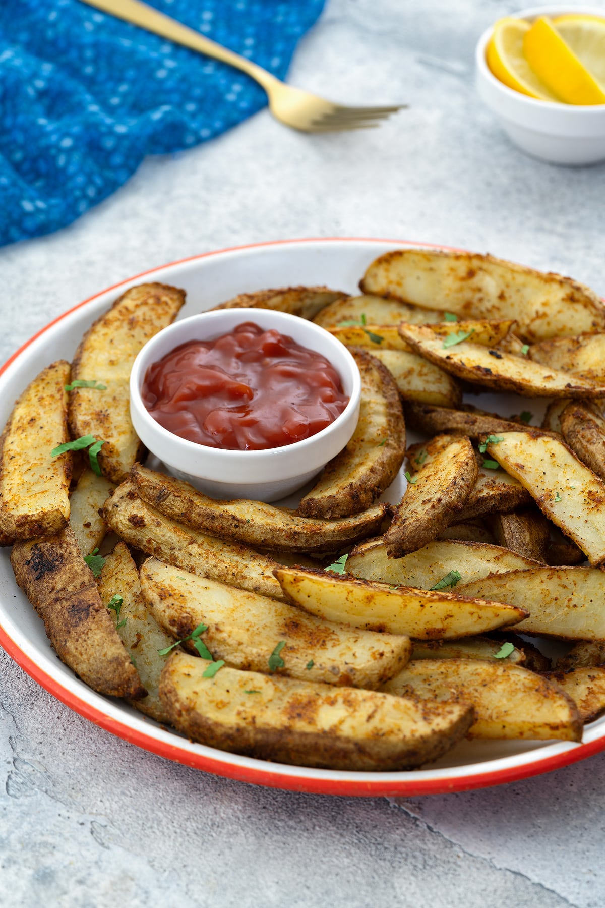 A plate of homemade potato wedges with a cup of ketchup on a white table. A blue towel, golden fork, and a small cup of lemon wedges are placed nearby.