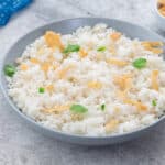 Coconut rice in a white bowl, garnished with coconut flakes and cilantro, on a white table. A blue towel and a cup of coconut flakes are placed in the top corners.