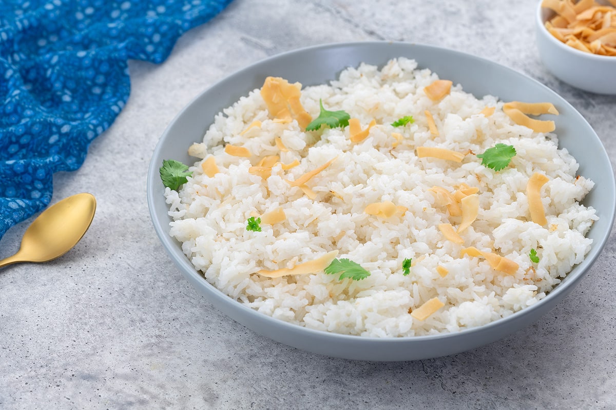 Coconut rice in a white bowl, garnished with coconut flakes and cilantro, placed on a white table. A blue towel, golden spoon, and a cup of coconut flakes are arranged nearby.