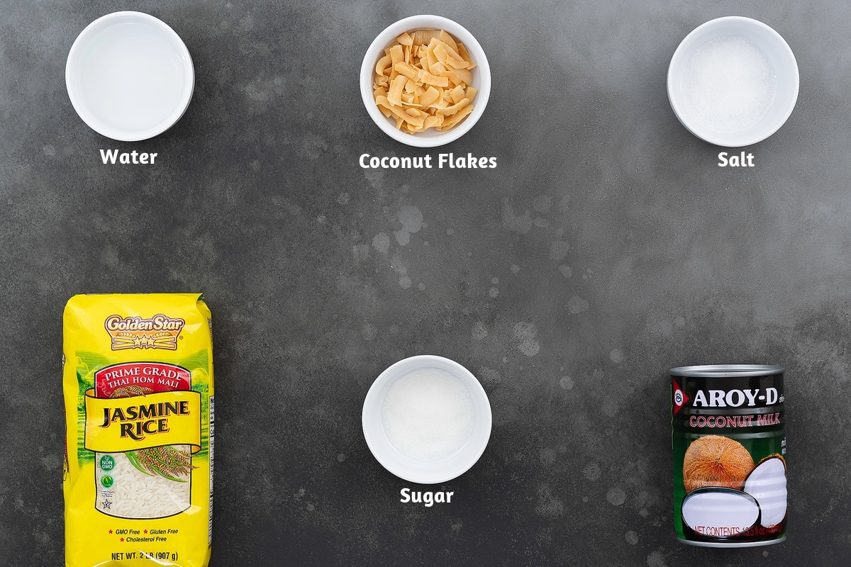 Ingredients for coconut rice recipe placed on a gray table, including water, coconut flakes, and salt on one side, and jasmine rice, sugar, and coconut milk.